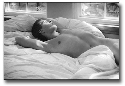 Man lying naked in a bed of white sheets with his body twisted toward the window looking out.