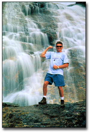 Terry Cyr standing at a waterfall in Montana flexing his muscle.