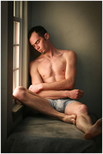 Nude male in gray shorts, looking down despondent, in morning window light